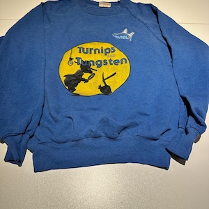 May include: Blue sweatshirt featuring the text "Turnips and Tungsten" in yellow, with a graphic of two figures. A small bird graphic is above the text. The sweatshirt has a crew neck and long sleeves.