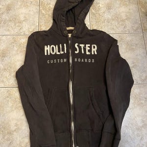 May include: A dark brown zip-up hoodie featuring a hood with metal eyelets and a front zipper. The chest displays the white text HOLLISTER and CUSTOM BOARDS in a distressed font. The garment includes two front pockets and ribbed cuffs.
