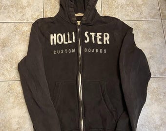 May include: A dark brown zip-up hoodie featuring a hood with metal eyelets and a front zipper. The chest displays the white text HOLLISTER and CUSTOM BOARDS in a distressed font. The garment includes two front pockets and ribbed cuffs.
