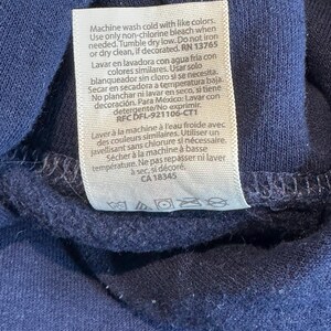 May include: A navy blue garment featuring a rectangular white care instruction label sewn onto the fabric. The label contains text in English, Spanish, and French, detailing washing, bleaching, drying, and ironing instructions with symbols.