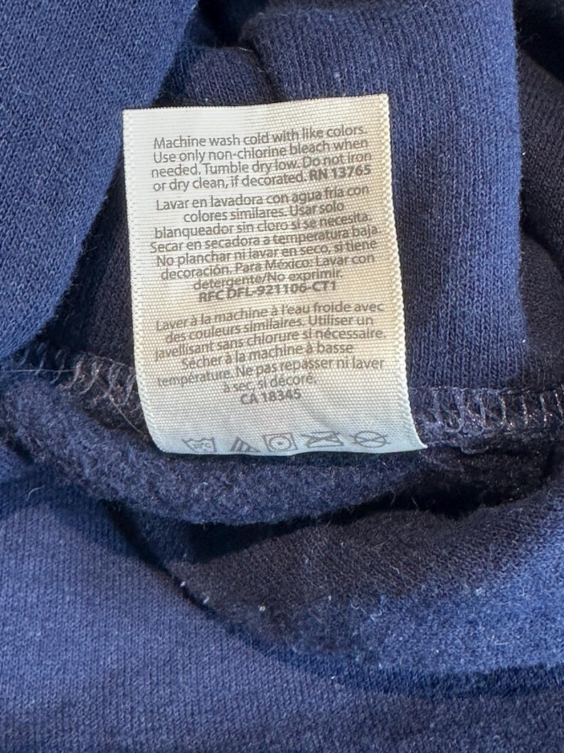 May include: A navy blue garment featuring a rectangular white care instruction label sewn onto the fabric. The label contains text in English, Spanish, and French, detailing washing, bleaching, drying, and ironing instructions with symbols.