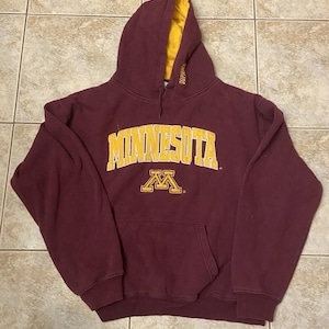 May include: Maroon hoodie with a gold-coloured interior lining. The word "MINNESOTA" is printed across the chest in gold block letters, with a gold "M" logo below. The hoodie has a front pocket and a drawstring hood.
