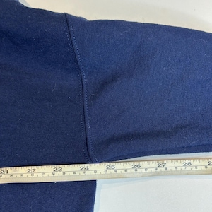 May include: A dark blue long-sleeved garment featuring a visible seam along the shoulder and arm. A metal ruler is positioned horizontally across the fabric, showing measurements from 21 inches to 28 inches along the top edge of the garment.