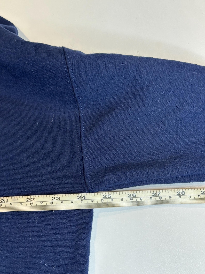 May include: A dark blue long-sleeved garment featuring a visible seam along the shoulder and arm. A metal ruler is positioned horizontally across the fabric, showing measurements from 21 inches to 28 inches along the top edge of the garment.
