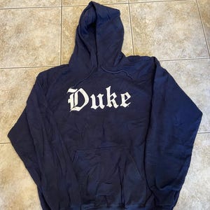 Può includere: Una felpa con cappuccio blu navy scuro con la scritta Duke stampata in un carattere gotico bianco sul petto. Il capo presenta una tasca a marsupio frontale, maniche lunghe con polsini a coste e un cordoncino abbinato sullo scollo.