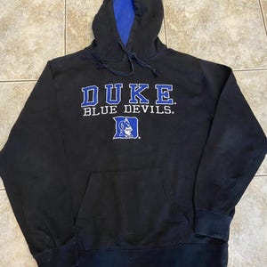 Może przedstawiać: Czarna bluza z kapturem z napisem "DUKE BLUE DEVILS" w białych i niebieskich literach. Wnętrze kaptura jest niebieskie. Widoczna jest przednia kieszeń i logo Duke Blue Devils.