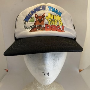 May include: A white and black baseball cap featuring a graphic of a brown dog with sharp teeth and a chain. The text on the front reads MEANER THAN A JUNK YARD DOG in blue, yellow, and red lettering. The cap has a black brim and a white front panel.