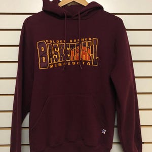 Könnte beinhalten: Ein kastanienbrauner Kapuzenpullover mit langen Ärmeln, einer Kängurutasche auf der Vorderseite und einer Kapuze mit Kordelzug. Auf der Brust befindet sich ein Aufdruck mit dem Text "GOLDEN GOPHERS BASKETBALL MINNESOTA" in gelben und orangefarbenen Buchstaben sowie einer Basketball-Grafik.