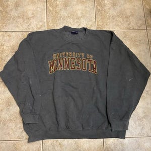 Puede incluir: Una sudadera de cuello redondo gris oscuro con el texto University of Minnesota impreso en color granate con un borde dorado en el pecho. La prenda de manga larga tiene cuello, puños y dobladillo acanalados, con marcas claras visibles.