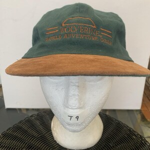 Puede incluir: Una gorra de béisbol de color verde oscuro con una visera de aspecto de ante marrón. La parte delantera presenta un texto bordado en naranja que dice WOLVERINE WORLD ADVENTURE GEAR con una línea curva sobre la palabra Wolverine y dos líneas horizontales.
