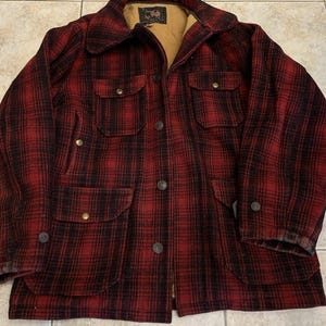 Puede incluir: Chaqueta a cuadros rojos y negros con cierre de botones, cuatro bolsillos delanteros y cuello. La chaqueta tiene forro beige y una etiqueta en el interior del cuello. Fabricada con un material grueso y cálido, ideal para el frío.