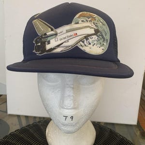 Könnte beinhalten: Eine marineblaue Baseballkappe mit einem grafischen Aufnäher eines weißen NASA-Space-Shuttles, das die Erde umkreist. Auf dem Shuttle stehen die Schriftzüge NASA und United States. Die Kappe hat ein strukturiertes Vorderteil und einen flachen Schirm.