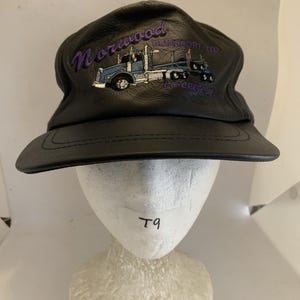Può includere: Un cappellino da baseball in pelle nera con un ricamo raffigurante un autoarticolato blu. Il testo "Norwood TRANSPORT LTD." e "LA CRETE" è cucito con filo viola sopra e sotto l'illustrazione del camion sul pannello frontale.