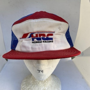 May include: A red, white, and blue trucker hat featuring a white front panel with the red and blue HRC Honda Racing logo printed in the center. The cap has a red brim, a white front crown, and blue mesh side and back panels for ventilation.