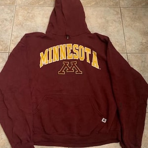 May include: A maroon hooded sweatshirt featuring the word MINNESOTA in yellow block letters with a yellow block M logo underneath. The hoodie includes a front kangaroo pocket and a small white Russell Athletic brand tag on the lower right side.