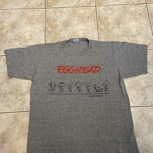 May include: A heather gray t-shirt with the word "EGGHEAD" in red block letters across the chest. Below the text is a black and white cartoon illustration of several characters. The shirt is laid flat on a tiled surface.