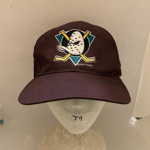 Op de afbeelding: Een donkerbruine honkbalpet met een geborduurd Mighty Ducks-logo op de voorkant. Het logo toont een wit eendenmasker met zwarte stippen, geplaatst over twee gekruiste gele hockeysticks op een groenblauwe achtergrond. De tekst Mighty Ducks is zichtbaar.
