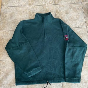 Pode incluir: Um suéter de fleece verde-escuro com zíper parcial, mangas compridas e bainha com cordão. Um pequeno emblema retangular com o texto "Downhill" e "LT" é visível na manga esquerda. A peça possui uma gola alta e uma textura macia.