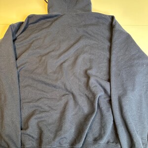 May include: A solid dark navy blue long-sleeve hooded sweatshirt featuring a drawstring hood, long sleeves with ribbed cuffs, and a ribbed hem. The garment is displayed from the back, showing a plain, unadorned fabric surface throughout the entire piece.