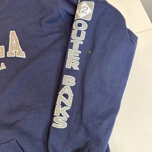 May include: A navy blue long-sleeve hooded sweatshirt featuring light gray text on the sleeve that reads "NC 12 OUTER BANKS" with a diamond-shaped graphic. The front of the garment displays partial white lettering, and the fabric has a smooth texture.