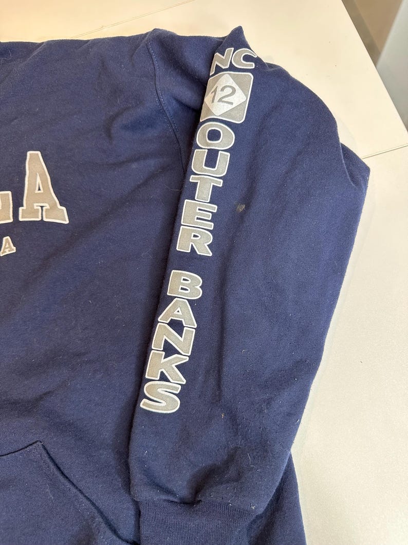 May include: A navy blue long-sleeve hooded sweatshirt featuring light gray text on the sleeve that reads "NC 12 OUTER BANKS" with a diamond-shaped graphic. The front of the garment displays partial white lettering, and the fabric has a smooth texture.