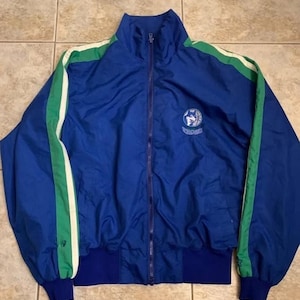 May include: A blue zip-up track jacket featuring a high collar, long sleeves with green and white side stripes, and a ribbed blue hem. A circular logo with a wolf graphic and the text Timberwolves is embroidered on the upper left chest area.