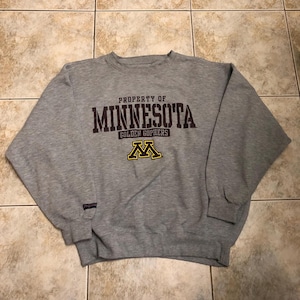 May include: A heather grey crewneck sweatshirt featuring the text PROPERTY OF MINNESOTA GOLDEN GOPHERS in dark brown and a yellow and brown M logo. A small black JanSport brand tag is visible on the right sleeve cuff of the long-sleeved garment.
