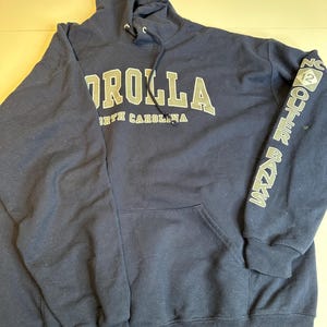 May include: A navy blue hooded sweatshirt featuring the text COROLLA NORTH CAROLINA in white and grey lettering on the chest. The left sleeve displays the text OUTER BANKS with a diamond-shaped NC 12 logo. It includes a front kangaroo pocket.