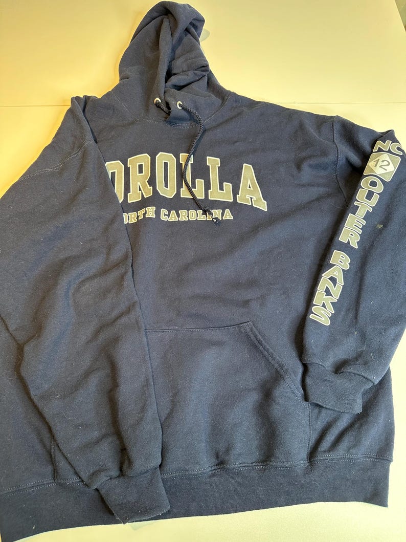 May include: A navy blue hooded sweatshirt featuring the text COROLLA NORTH CAROLINA in white and grey lettering on the chest. The left sleeve displays the text OUTER BANKS with a diamond-shaped NC 12 logo. It includes a front kangaroo pocket.
