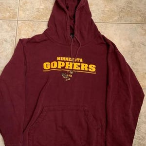 May include: A maroon hooded sweatshirt featuring the text MINNESOTA GOPHERS in yellow lettering across the chest. Below the text is a small graphic of a gopher mascot. The hoodie includes a front kangaroo pocket and matching maroon drawstrings.