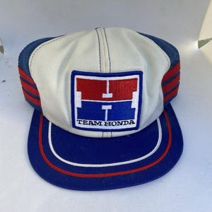 以下が含まれることがあります： 白いフロントパネルに、赤と青のグラフィックロゴと「TEAM HONDA」の文字が入った長方形のパッチが付いたトラッカーキャップ。青いツバには赤と白のストライプがあり、青いメッシュのサイドには赤いストライプが施されています。