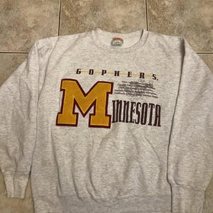 May include: A light grey crewneck sweatshirt featuring a large yellow and maroon letter M, the text GOPHERS and MINNESOTA, and additional printed details including team colours, address, and stadium names in maroon and black lettering.