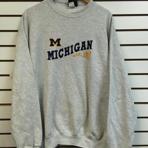 Può includere: Una felpa girocollo grigio chiaro con il testo ricamato MICHIGAN in blu scuro, una lettera M blu scuro e il testo giallo ricamato EST. 1817. Il capo presenta maniche lunghe con polsini a coste e uno scollo a coste, appeso su una gruccia.