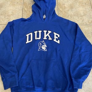 Può includere: Felpa con cappuccio blu reale con la scritta "DUKE" a lettere bianche sul petto. Sotto, una grafica bianca di un Blue Devil. La felpa ha una tasca frontale e un cappuccio con coulisse.