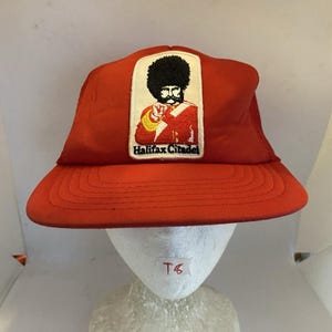 May include: A bright red baseball cap featuring a rectangular embroidered patch on the front. The patch depicts a soldier in a red uniform and a tall black bearskin hat, with the text Halifax Citadel printed in black lettering below the figure.