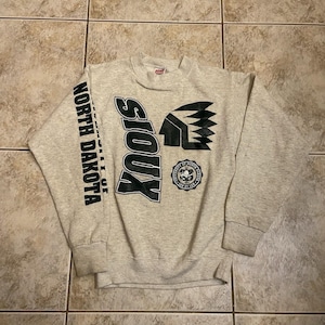 May include: A heather gray crewneck sweatshirt featuring black screen-printed graphics. The front displays the word SIOUX, a stylized Native American headdress icon, and a circular University of North Dakota seal. The right sleeve reads NORTH DAKOTA.