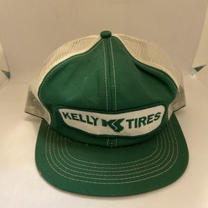 May include: A green and white trucker hat featuring a mesh back and a solid green front panel. A white rectangular patch is centered on the front with the green text Kelly Tires and a stylized K logo. The brim is green with white stitching.