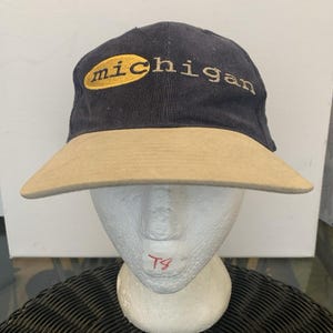 May include: A navy blue baseball cap with a tan brim and the word "michigan" embroidered in gold thread on the front. The text "michigan" is partially enclosed within a gold oval shape. The cap features a structured crown and a curved visor.