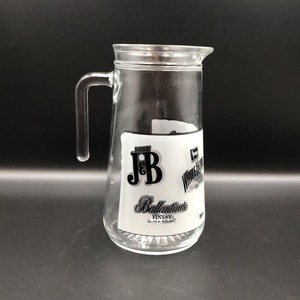 Puede incluir: Una jarra de vidrio transparente con asa. La jarra tiene una etiqueta con el texto "Rare J&B" y "Ballantine's Finest Scotch Whisky".