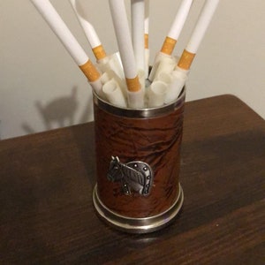 Op de afbeelding: Een zilveren sigarettenhouder met een leren basis en een paardenontwerp. De houder heeft een draaibaar bovendeel met meerdere sleuven voor sigaretten.