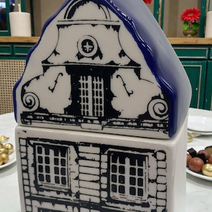 Op de afbeelding: Een witte keramische huisvormige container met een blauw dak. De container heeft zwarte architectonische details, waaronder ramen en decoratieve elementen. Het ontwerp doet denken aan traditionele Nederlandse huizen.