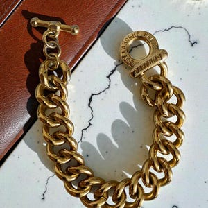 Peut inclure: Un bracelet chaîne doré avec fermoir à bascule. Le bracelet présente des maillons épais et arrondis et un fermoir circulaire gravé "Guy Laroche Paris". Le bracelet est présenté sur une surface blanche et grise avec un fond en cuir marron.