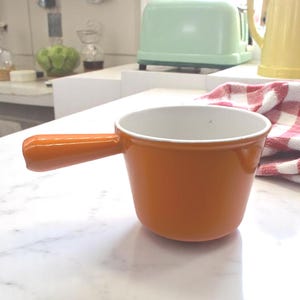 May include: Orange enamel cast iron saucepan with a white interior and a single handle. The saucepan is sitting on a white marble countertop.