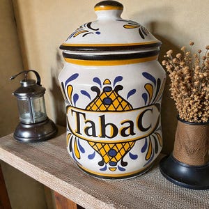 Peut inclure: Un pot en céramique blanc avec un couvercle, portant l'inscription "Tabac". Le pot est orné de motifs floraux jaunes et bleus et d'accents noirs. Une petite lanterne et un vase décoratif sont sur l'étagère.