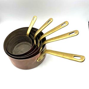 May include: Set of five copper saucepans with brass handles. The saucepans are nested inside each other, with the largest saucepan at the bottom and the smallest saucepan at the top.