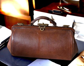 May include: A brown leather doctor's bag with brass hardware and a top handle. The bag is sitting on a dark surface.