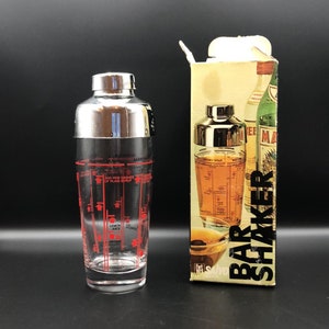 Peut inclure: Un shaker à cocktail en argent et en verre avec des marques rouges et des mesures pour différentes boissons. Le shaker est à côté d'une boîte sur laquelle est imprimé "Bar Shaker".