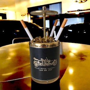 Peut inclure: Un porte-cigarettes noir et or avec un logo Rolls-Royce Silver Ghost. Le porte-cigarettes a un dessus rond et une base cylindrique. Il y a trois cigarettes dans le porte-cigarettes.