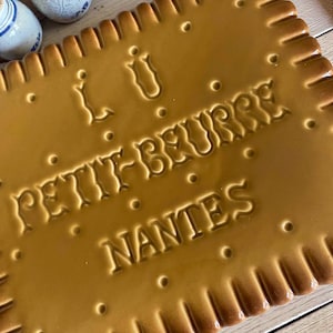 Peut inclure: Un plat de service rectangulaire en céramique en forme de biscuit, avec un glaçage marron. Les mots "Petit-Beurre Nantes" sont en relief sur la surface. Le plat a un bord festonné et de petits détails en forme de points.
