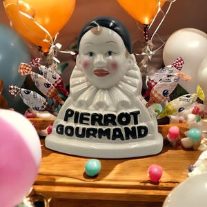 Puede incluir: Una figurita de cerámica blanca de un Pierrot payaso con un sombrero negro y un cuello con volantes. La figurita sostiene una variedad de caramelos coloridos envueltos en celofán. El texto "PIERROT GOURMAND" está escrito en la base de la figurita.